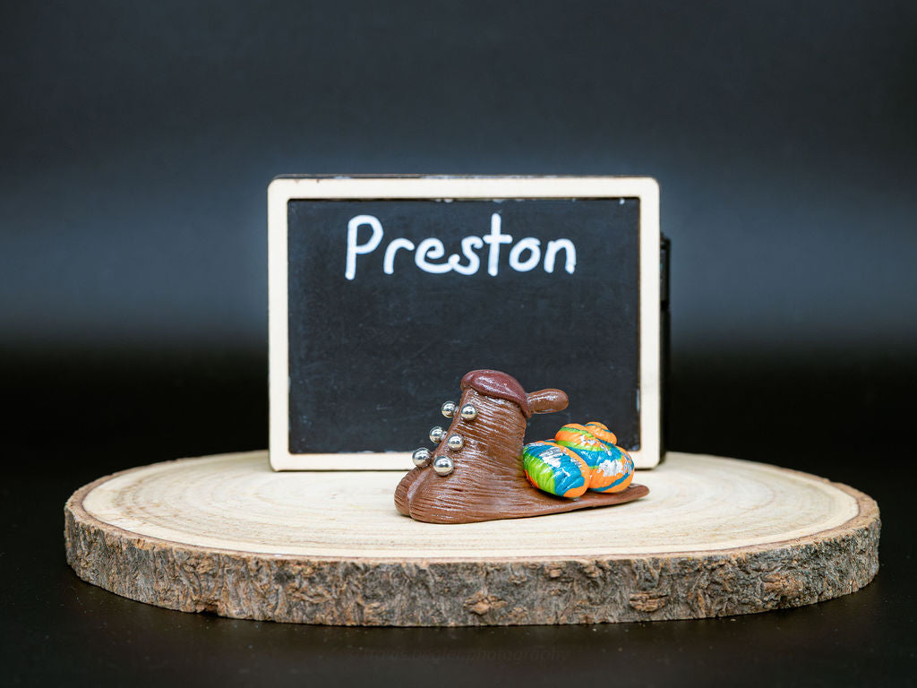 Small snail with piercings and a decorative shell on a wooden platform in front of a chalkboard with the name 'Preston' written on it
